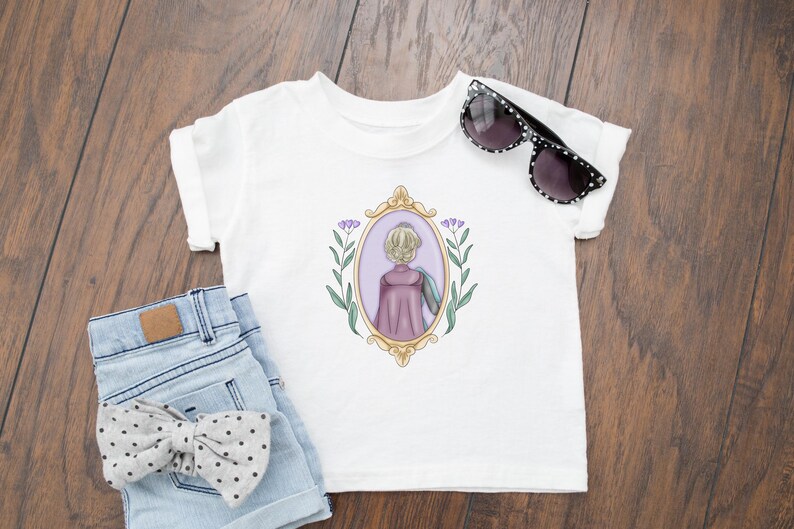 Peut inclure: T-shirt blanc avec une illustration violette et dor&eacute;e d'une femme aux cheveux blonds dans un cadre avec des fleurs violettes. La femme porte une cape violette et regarde loin du spectateur.