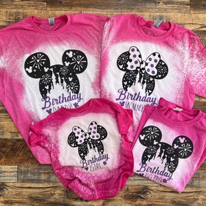 Peut inclure: Quatre t-shirts roses avec des motifs &eacute;clabouss&eacute;s de javel repr&eacute;sentant une silhouette noire d'oreilles de Mickey Mouse avec des feux d'artifice et le texte "Birthday Daddy", "Birthday Mommy", "Birthday Girl" et "Birthday Big Bro".