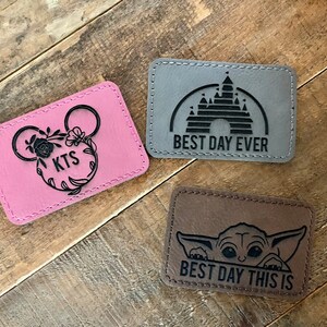 May include: Three leather patches with different designs. The first patch is pink with a black Mickey Mouse head and the text "KTS". The second patch is gray with a black silhouette of Cinderella's castle and the text "BEST DAY EVER". The third patch is brown with a black silhouette of Baby Yoda and the text "BEST DAY THIS IS".