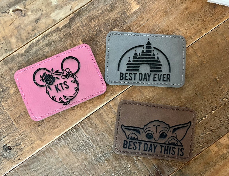 May include: Three leather patches with different designs. The first patch is pink with a black Mickey Mouse head and the text "KTS". The second patch is gray with a black silhouette of Cinderella's castle and the text "BEST DAY EVER". The third patch is brown with a black silhouette of Baby Yoda and the text "BEST DAY THIS IS".