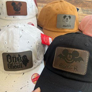 May include: Five different baseball caps with personalized patches. The patches are brown leather with a Mickey Mouse design and a name. The caps are black, white, pink, and brown.