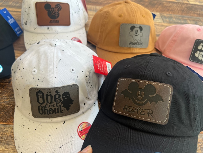 May include: Five different baseball caps with personalized patches. The patches are brown leather with a Mickey Mouse design and a name. The caps are black, white, pink, and brown.