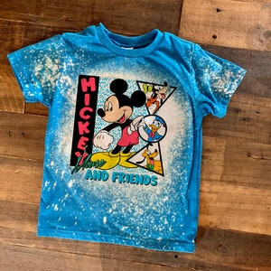 Puede incluir: Una camiseta azul con un dise&ntilde;o salpicado de lej&iacute;a con Mickey Mouse y sus amigos. La camiseta tiene el texto "Mickey Mouse and Friends" impreso.