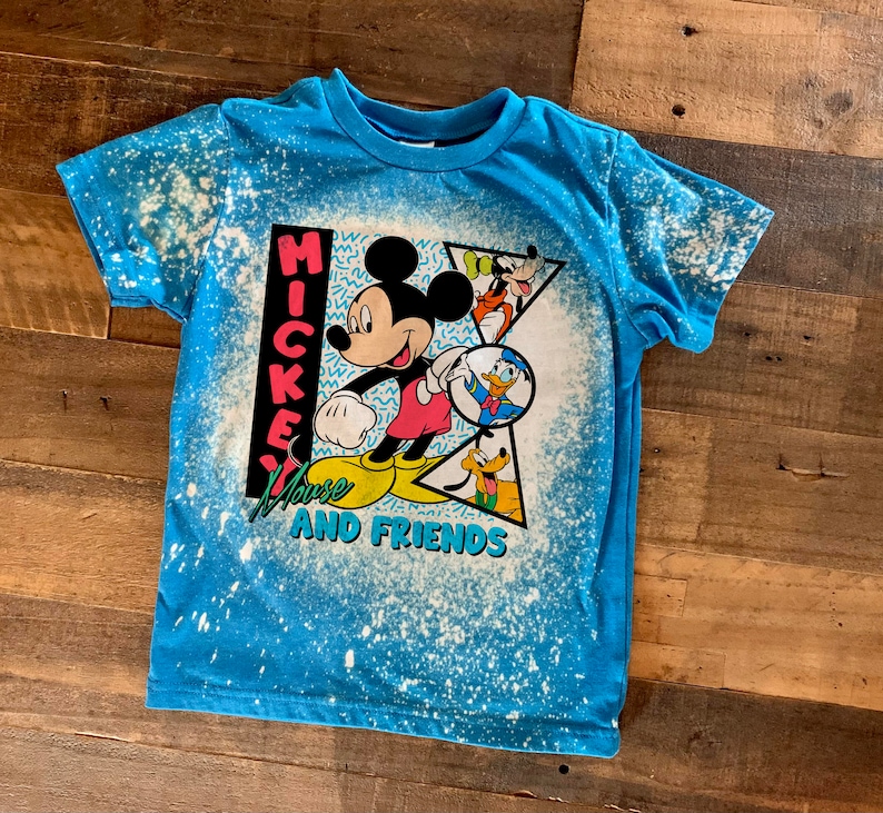 Puede incluir: Una camiseta azul con un dise&ntilde;o salpicado de lej&iacute;a con Mickey Mouse y sus amigos. La camiseta tiene el texto "Mickey Mouse and Friends" impreso.