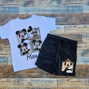 May include: White t-shirt with a cartoon image of Mickey Mouse in a safari hat. The shirt has the name "Max" printed on it. Black shorts with a cartoon image of Mickey Mouse in a safari hat on the right leg.