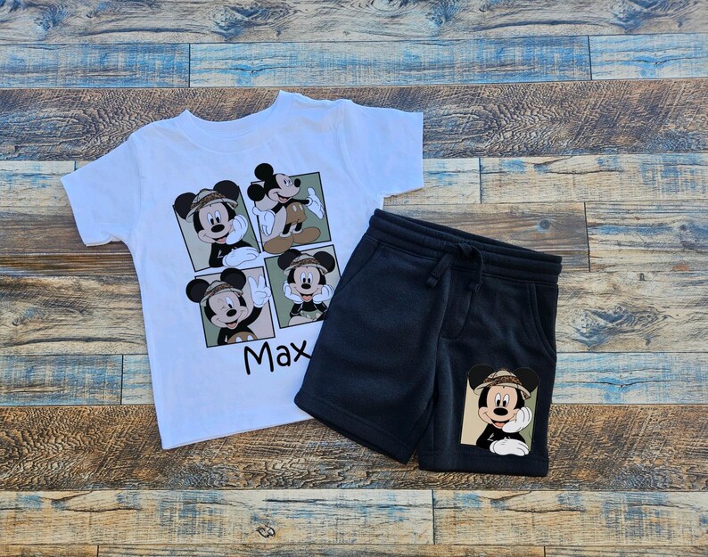 May include: White t-shirt with a cartoon image of Mickey Mouse in a safari hat. The shirt has the name "Max" printed on it. Black shorts with a cartoon image of Mickey Mouse in a safari hat on the right leg.