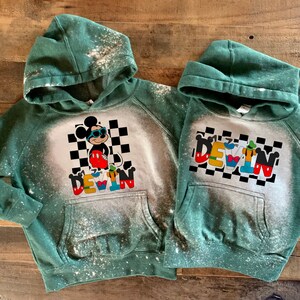 May include: Two green hooded sweatshirts with bleach-splattered designs. One features Mickey Mouse wearing sunglasses and the name "DEVIN" in colorful letters. The other features the name "DEVIN" spelled out with different Disney characters.