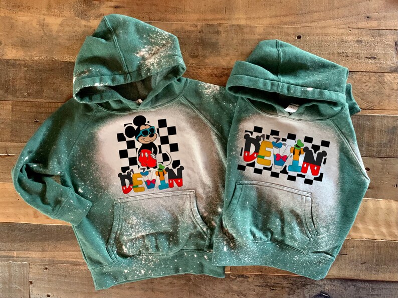 May include: Two green hooded sweatshirts with bleach-splattered designs. One features Mickey Mouse wearing sunglasses and the name "DEVIN" in colorful letters. The other features the name "DEVIN" spelled out with different Disney characters.