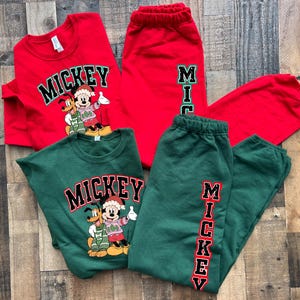 May include: Two sets of matching Christmas-themed clothing featuring Mickey Mouse and Pluto. One set is red with green accents and the other is green with red accents. Both sets include a long-sleeved shirt with the word "MICKEY" printed on the front and sweatpants with the word "MICKEY" printed down the side.