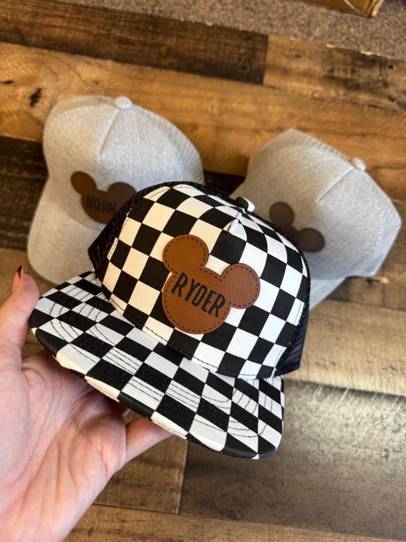 May include: A black and white chequered trucker hat with the name "Ryder" on a brown leather patch. Two additional grey hats with brown Mickey Mouse-shaped patches are also visible. The hats are on a wooden surface.