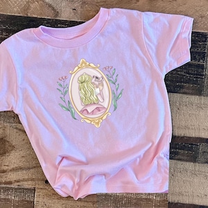 Peut inclure: Un t-shirt rose avec un motif repr&eacute;sentant une femme dans un cadre dor&eacute; avec des fleurs. La femme tient un petit oiseau dans sa main.