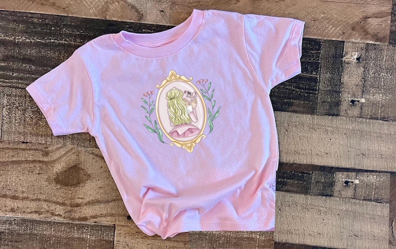 Peut inclure: Un t-shirt rose avec un motif repr&eacute;sentant une femme dans un cadre dor&eacute; avec des fleurs. La femme tient un petit oiseau dans sa main.