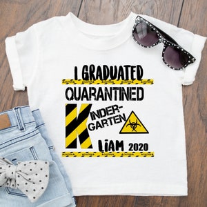 May include: White t-shirt with black text that reads "I Graduated Quarantined Kindergarten Liam 2020". The text is surrounded by yellow and black caution tape. A black and yellow letter K is in the center of the shirt with a biohazard symbol in a yellow triangle.