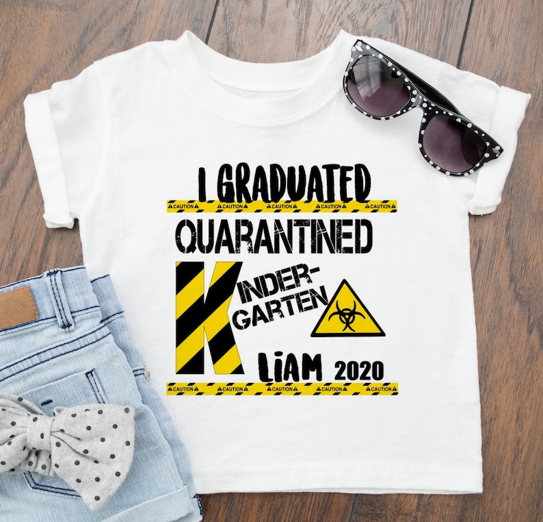 May include: White t-shirt with black text that reads "I Graduated Quarantined Kindergarten Liam 2020". The text is surrounded by yellow and black caution tape. A black and yellow letter K is in the center of the shirt with a biohazard symbol in a yellow triangle.