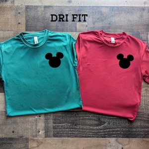 May include: Two short-sleeved t-shirts, one teal and one red, each featuring a black Mickey Mouse head graphic on the chest. The word "DRI FIT" is printed in black at the top. The shirts are displayed on a wooden surface.