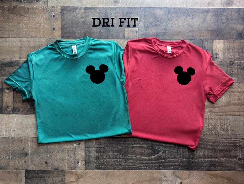 May include: Two short-sleeved t-shirts, one teal and one red, each featuring a black Mickey Mouse head graphic on the chest. The word "DRI FIT" is printed in black at the top. The shirts are displayed on a wooden surface.