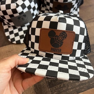 K&ouml;nnte beinhalten: Schwarz-wei&szlig; karierte Trucker-Caps mit einem braunen Lederpatch mit einem von Mickey Mouse inspirierten Design. Auf einer Kappe steht der Name "PARKER" auf dem Patch. Die Kappen haben eine schwarze Mesh-R&uuml;ckseite und einen gebogenen Schirm.