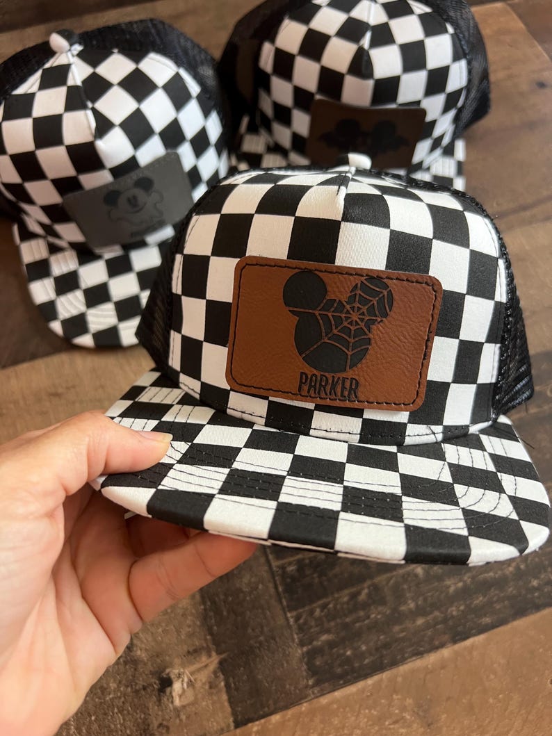 K&ouml;nnte beinhalten: Schwarz-wei&szlig; karierte Trucker-Caps mit einem braunen Lederpatch mit einem von Mickey Mouse inspirierten Design. Auf einer Kappe steht der Name "PARKER" auf dem Patch. Die Kappen haben eine schwarze Mesh-R&uuml;ckseite und einen gebogenen Schirm.