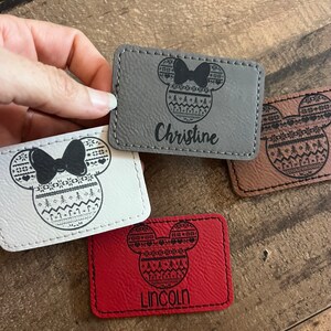 May include: Three personalized leather keychains with a Mickey Mouse head design featuring a Christmas sweater pattern. The keychains are in gray, white, and red. The names "Christine", "Lincoln" are engraved on the keychains.
