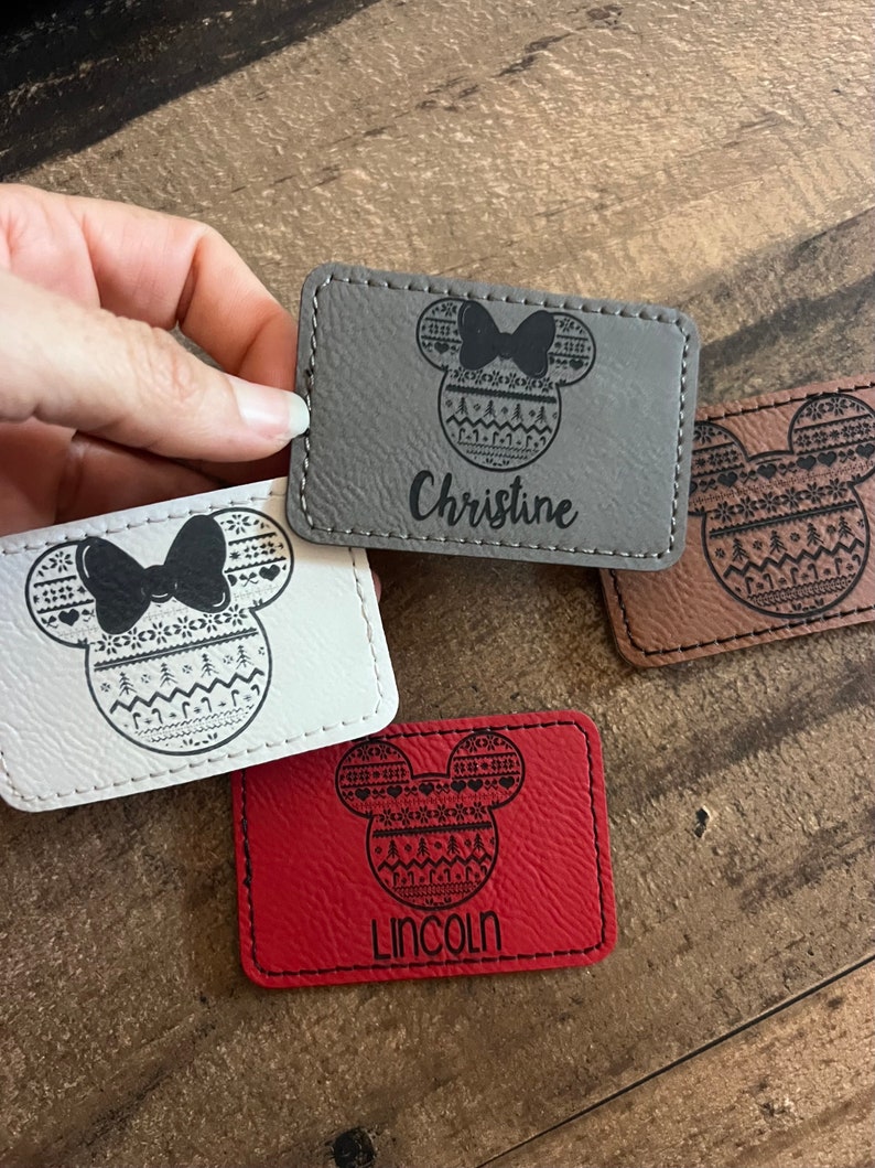 May include: Three personalized leather keychains with a Mickey Mouse head design featuring a Christmas sweater pattern. The keychains are in gray, white, and red. The names "Christine", "Lincoln" are engraved on the keychains.