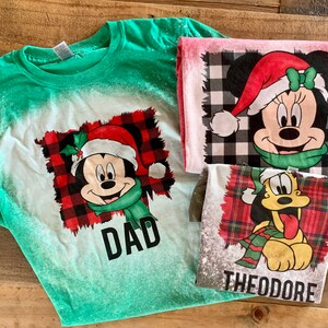 May include: Three Christmas-themed t-shirts featuring Mickey Mouse, Minnie Mouse, and Pluto. The shirts are green, pink, and gray with a white bleach effect. The shirts have a red and black plaid pattern and the text "DAD" and "THEODORE" on them.