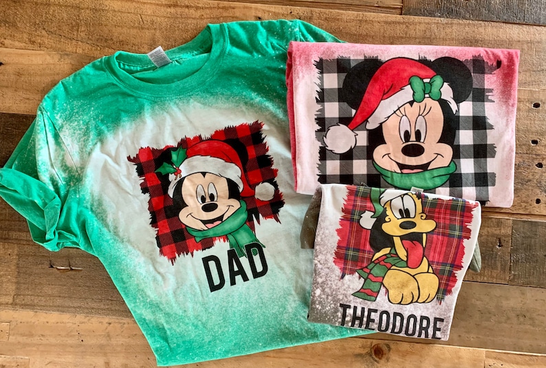 May include: Three Christmas-themed t-shirts featuring Mickey Mouse, Minnie Mouse, and Pluto. The shirts are green, pink, and gray with a white bleach effect. The shirts have a red and black plaid pattern and the text "DAD" and "THEODORE" on them.