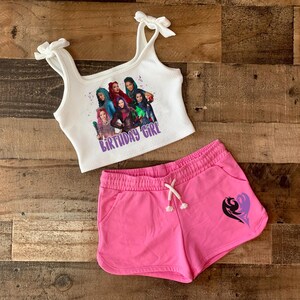 May include: White tank top with a graphic of characters from the Disney movie Descendants. The text "Birthday Girl" is printed on the tank top. The tank top is paired with pink shorts that have a purple heart design.