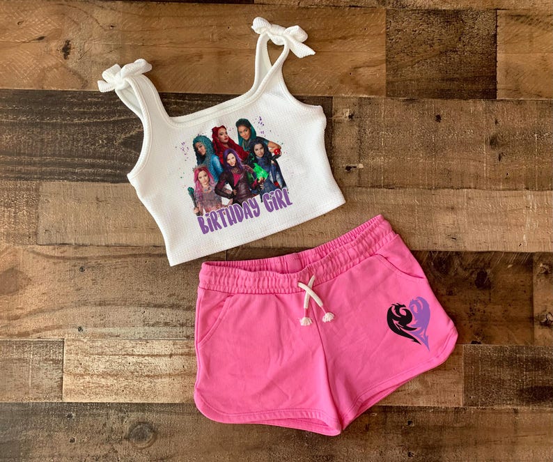 May include: White tank top with a graphic of characters from the Disney movie Descendants. The text "Birthday Girl" is printed on the tank top. The tank top is paired with pink shorts that have a purple heart design.
