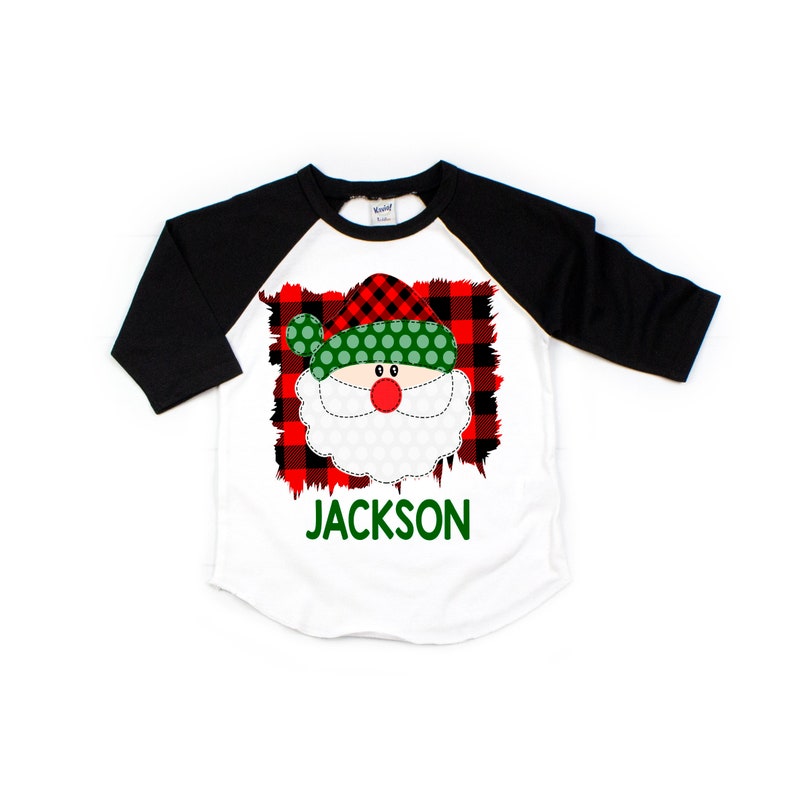 May include: A white and black baseball tee shirt with a red and green plaid Santa Claus face graphic. The text "JACKSON" is printed in green below the graphic.