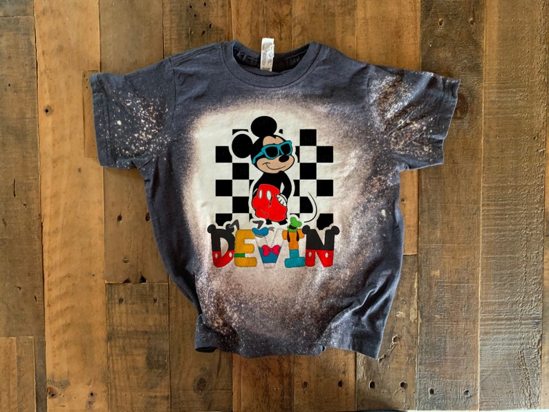 May include: A dark blue t-shirt with a bleach-splattered design featuring Mickey Mouse wearing sunglasses and a checkered background. The name "DEVIN" is spelled out below Mickey Mouse in colorful letters.