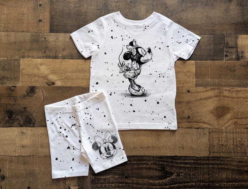 May include: A white t-shirt and shorts set with a black and white print of Mickey Mouse. The shirt has a crew neck and short sleeves. The shorts have an elastic waistband.
