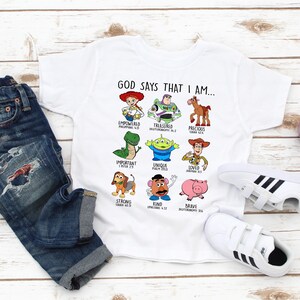 May include: White t-shirt with a graphic of Toy Story characters and the text "GOD SAYS THAT I AM..." with bible verses underneath each character. The shirt is on a white wooden surface with blue jeans and white sneakers.