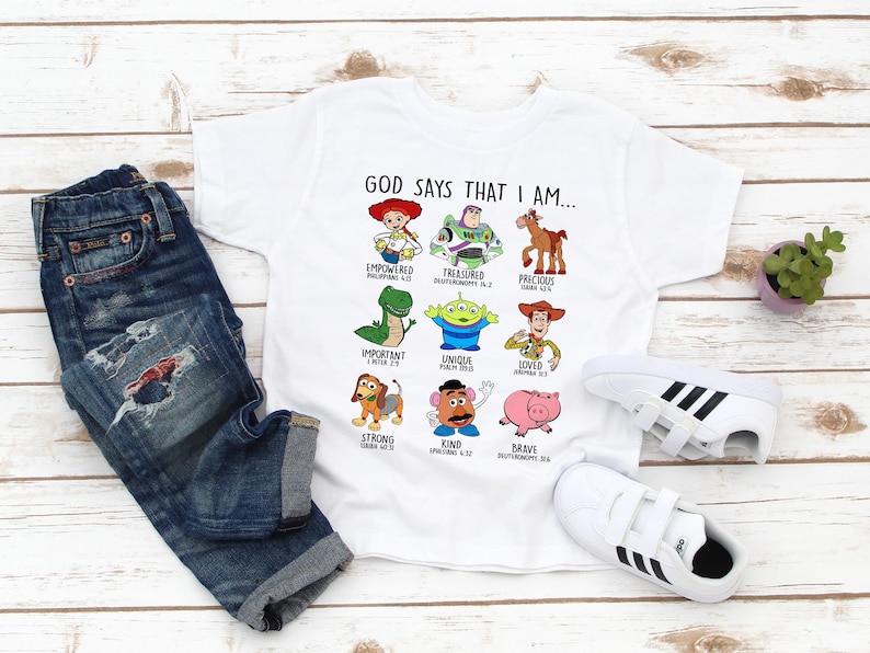 May include: White t-shirt with a graphic of Toy Story characters and the text "GOD SAYS THAT I AM..." with bible verses underneath each character. The shirt is on a white wooden surface with blue jeans and white sneakers.