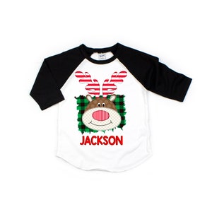 May include: A white and black baseball tee shirt with a red and white reindeer face graphic. The reindeer has a red nose and green and black plaid background. The text "JACKSON" is printed in red below the reindeer.