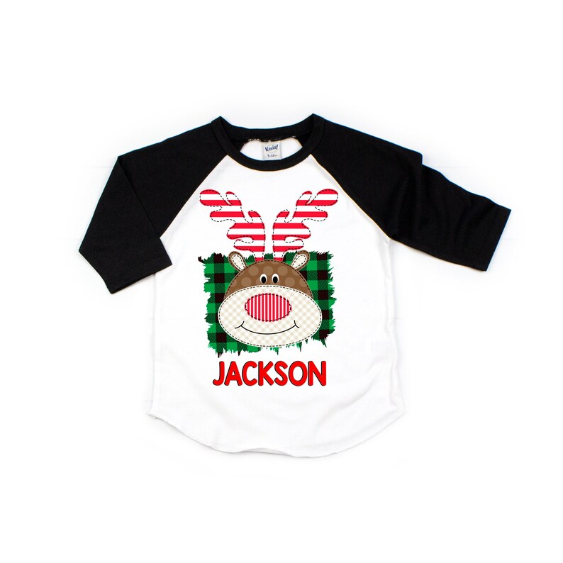 May include: A white and black baseball tee shirt with a red and white reindeer face graphic. The reindeer has a red nose and green and black plaid background. The text "JACKSON" is printed in red below the reindeer.