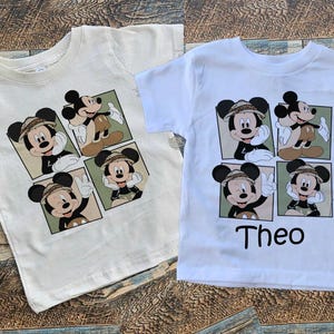 May include: Two white t-shirts with a cartoon image of Mickey Mouse in different poses. The shirts have a green and brown background. The text "Theo" is printed on the bottom of one of the shirts.