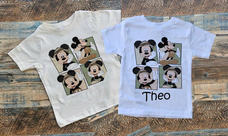 May include: Two white t-shirts with a cartoon image of Mickey Mouse in different poses. The shirts have a green and brown background. The text "Theo" is printed on the bottom of one of the shirts.