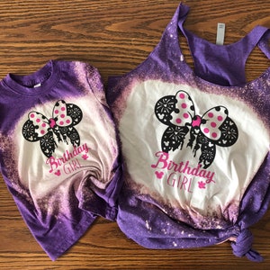 Peut inclure: Deux t-shirts en coton blanchis, de couleur violette et blanche, avec un n&oelig;ud Minnie Mouse noir et blanc avec des pois roses et le texte "Birthday Girl".
