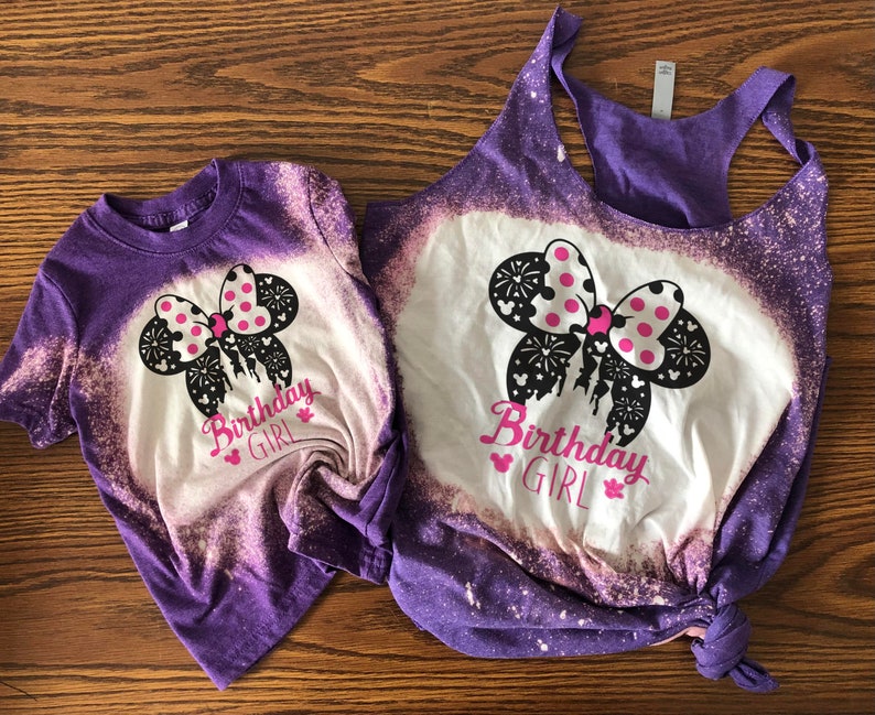 Peut inclure: Deux t-shirts en coton blanchis, de couleur violette et blanche, avec un n&oelig;ud Minnie Mouse noir et blanc avec des pois roses et le texte "Birthday Girl".