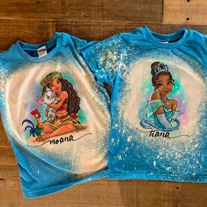 Peut inclure: Deux t-shirts bleus avec des motifs &eacute;clabouss&eacute;s de javel. L'un repr&eacute;sente Moana avec Pua et un coq, l'autre repr&eacute;sente Tiana. Les deux t-shirts portent le nom du personnage imprim&eacute; sous l'image.