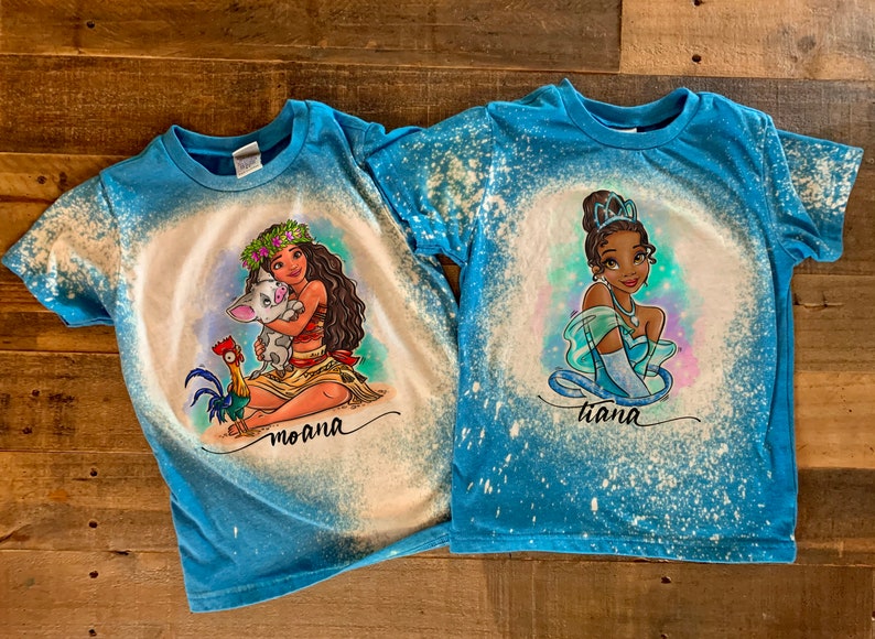 Peut inclure: Deux t-shirts bleus avec des motifs &eacute;clabouss&eacute;s de javel. L'un repr&eacute;sente Moana avec Pua et un coq, l'autre repr&eacute;sente Tiana. Les deux t-shirts portent le nom du personnage imprim&eacute; sous l'image.