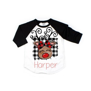 May include: A white and black baseball tee shirt with black sleeves. The shirt features a cartoon reindeer with a red bow and a red nose. The text "Harper" is printed in pink below the reindeer.