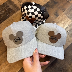 May include: Three baseball caps. Two grey caps have a brown Mickey Mouse head patch with the names "MAX" and "NOAH". The third cap has a black and white chequered pattern with a black mesh back and a brown patch that says "RYDER".