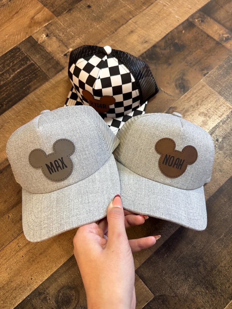 May include: Three baseball caps. Two grey caps have a brown Mickey Mouse head patch with the names "MAX" and "NOAH". The third cap has a black and white chequered pattern with a black mesh back and a brown patch that says "RYDER".