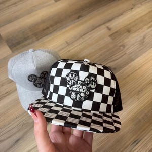 May include: Two baseball caps. One is grey with a black Mickey Mouse design. The other is black and white chequered with a Star Wars Mickey Mouse design. The Star Wars design includes characters and the words "Star Wars".