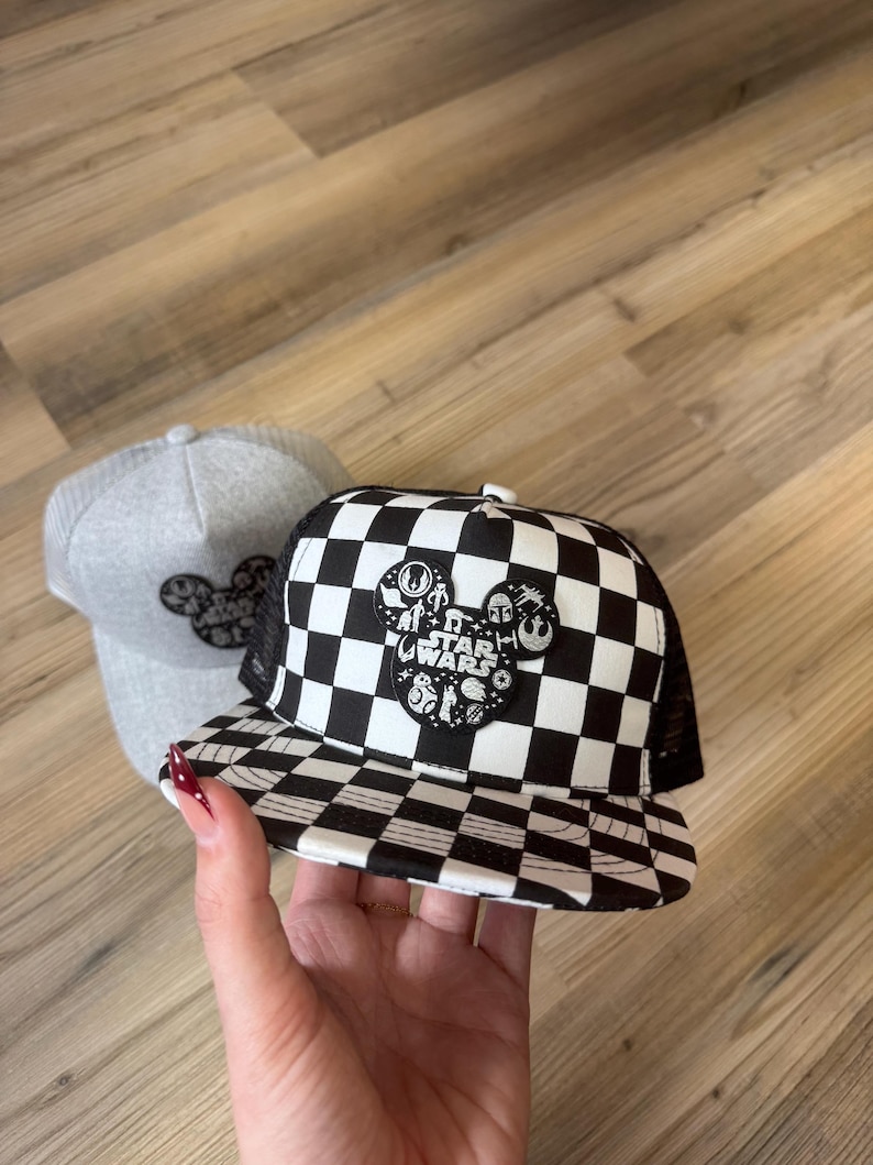 May include: Two baseball caps. One is grey with a black Mickey Mouse design. The other is black and white chequered with a Star Wars Mickey Mouse design. The Star Wars design includes characters and the words "Star Wars".