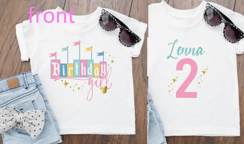 May include: Two white t-shirts with a pink and gold design. The first shirt has a castle silhouette with the text "Birthday girl" and a gold Mickey Mouse head. The second shirt has the name "Lenna" and the number "2" in pink with gold stars.