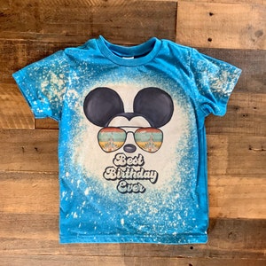 May include: A blue t-shirt with a white bleach splattered design. The design features a cartoon Mickey Mouse wearing sunglasses with a castle reflection. The text "Best Birthday Ever" is below the sunglasses.