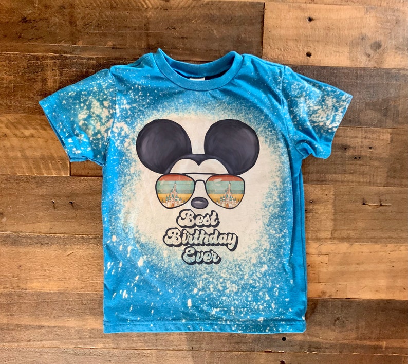 May include: A blue t-shirt with a white bleach splattered design. The design features a cartoon Mickey Mouse wearing sunglasses with a castle reflection. The text "Best Birthday Ever" is below the sunglasses.