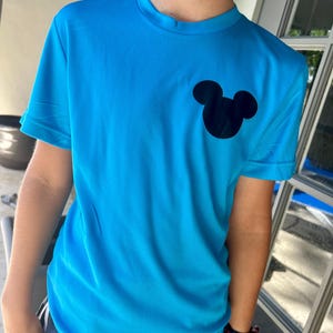 May include: A bright blue t-shirt featuring a black Mickey Mouse silhouette on the chest. The short-sleeved shirt is made of a lightweight, possibly athletic material. The shirt is a solid color with a crew neck.